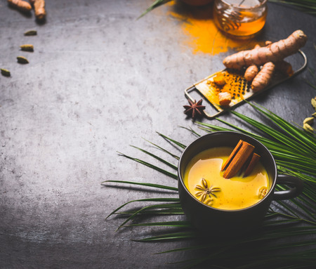 Black mug of vegan golden turmeric milk with spices on dark background with ingredients and palm leaves. Healthy hot drink. Immune boosting remedy , detox and dieting conceptの写真素材