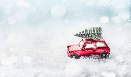 Christmas tree on roof of red retro car toy in snow through snowy winter wonderland with snowfall. Creative Christmas holiday concept. Copy space for your greeting and design. Front viewの写真素材