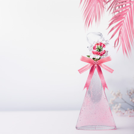 Stylized cosmetic glass bottle with pink ribbon and flowers and hanged palm leaves standing on white gray background with copy space. Skin care and beauty conceptの写真素材