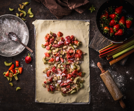 Sheet of dough with sliced rhubarb and strawberries. Strudel pie preparation on rustic kitchen table, top view. Seasonal baking. Organic food. Step by stepの写真素材