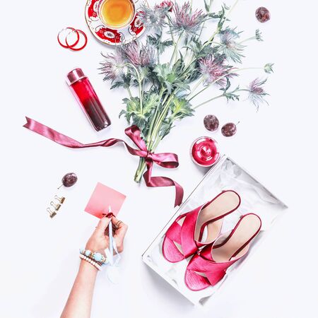 Woman hand writes a note on blank pink paper on white desk with high heels in box, cosmetics, perfume, flowers bunch, cakes cup of tea and bijou on white desk background. Casual woman beauty blogの写真素材
