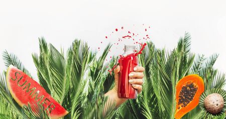 Female hand with red drink bottle: juice or smoothie leaned out of jungle  with fruits , coconut and water melon with text summer at white background. Healthy lifestyle. Summer mood. Splash.の写真素材