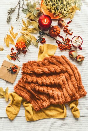 Female autumn outfit with sweater and  faux fur poncho on white blanket with fall leaves, pumpkins ,candle and mug with cappuccino , top view. Flat lay. Copy space. Autumn lifestyle mood. Cozy homeの写真素材