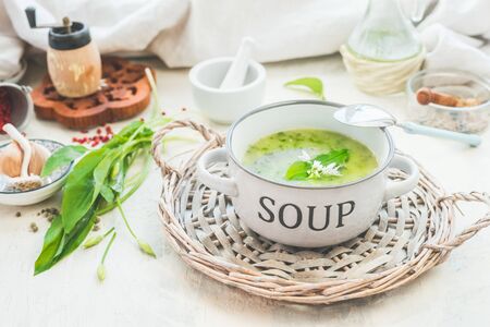 Green wild garlic soup in bowl with spoon on light table with ingredients. Close up. Seasonal home...