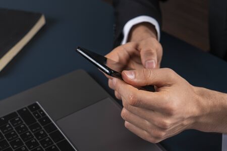 Close up of businessman hands with smartphone in blurred office with dark blue table and laptop....