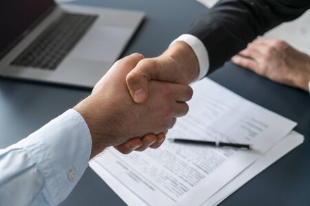 Close up of two businessmen shaking hands in blurred office with computer and contract on table....