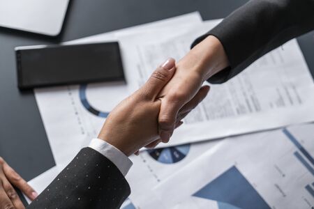 Top view of two businesswomen shaking hands at blurred office table with graphs. Concept of partnership and collaborationの写真素材