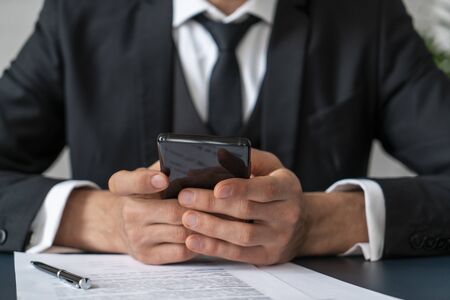 Front view of unrecognizable businessman in dark suit holding smartphone while sitting at office table with contract and pen. Focus on the hands. Blurred backgroundの写真素材