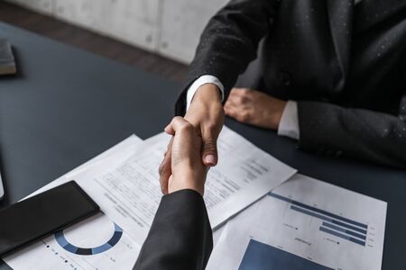 Top view of two businesswomen shaking hands at blurred office table with business reports and...