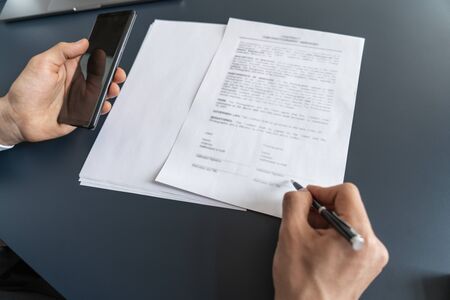 Top view of businessman hands signing documents and holding smartphone at blurred office table....