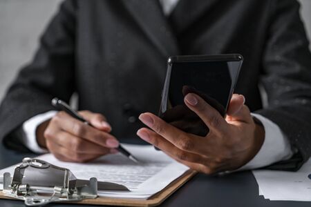 Close up of businesswoman looking at smartphone and working with document on clipboard at blurred...