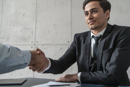 Confident and calm young businessman with dark hair shaking hands with his partner in loft office. Concept of partnership and employmentの写真素材