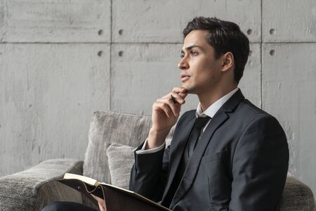 Side view of thoughtful and calm young businessman writing in his notebook sitting in gray armchair in loft office. Concept of business planningの写真素材