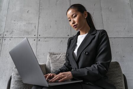 Concentrated young Asian businesswoman typing on laptop while sitting in armchair in loft office. Concept of business lifestyle and freelance jobの写真素材