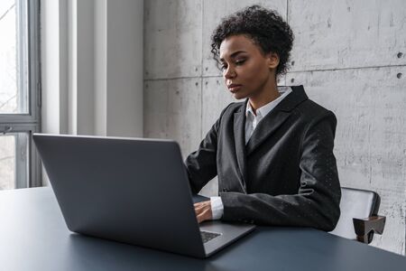 Gorgeous and serious young African woman in business suit working with laptop in loft office. Concept of business lifestyle and educationの写真素材