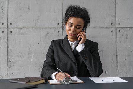 Portrait of young African American businesswoman talking on smartphone and taking notes in loft office. Concept of business communicationの写真素材