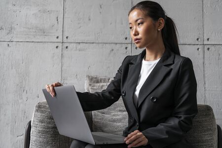 Concentrated young Asian woman in dark suit sitting with laptop in soft gray armchair in loft...