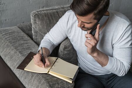 Top view of handsome bearded businessman in casual clothes talking on smartphone and writing in armchair in loft office. Concept of freelance and schedulingの写真素材