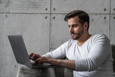 Serious young businessman in casual clothes typing on laptop sitting in armchair in loft office or...