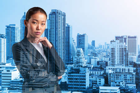 Attractive young Asian business woman in suit with hand on chin thinking how to succeed, new career opportunities, MBA. Bangkok on background. Double exposure.の写真素材
