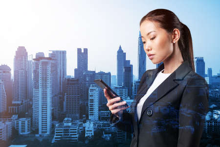 Prosperous astonishing young Asian business woman in suit texting phone the details of corporate meeting. Bangkok cityscape. The concept of business communication. Double exposure.の写真素材