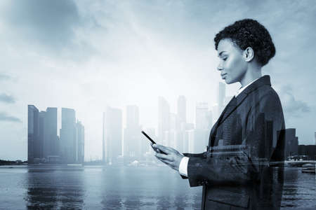 Attractive black African American business woman in suit using smart phone and thinking how to tackle the problem, new career opportunities, MBA. Singapore on background. Double exposure.の写真素材