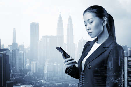 Prosperous astonishing young Asian business woman in suit texting phone the details of corporate meeting. Kuala Lumpur cityscape. The concept of business communication. Double exposure.の写真素材
