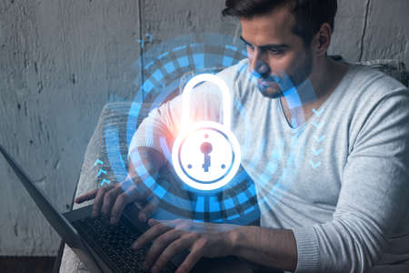 A serious caucasian programmer in casual wears, using laptop to develop a new security system to protect data. Double exposure. IT lock hologram icon.の写真素材
