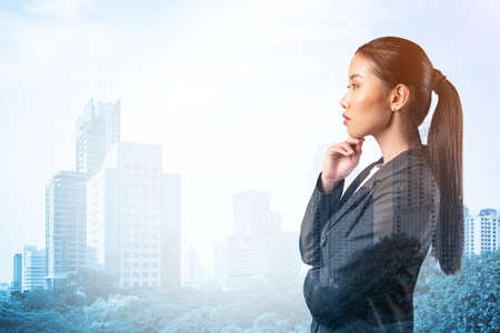 Attractive young Asian business woman in suit with hand on chin thinking how to succeed, new career opportunities, MBA. Bangkok on background. Double exposure.の写真素材