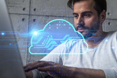 A serious caucasian man in casual wears, typing laptop to create a protection software to store confidential information. Double exposure. Hologram of an IT cloud.の写真素材