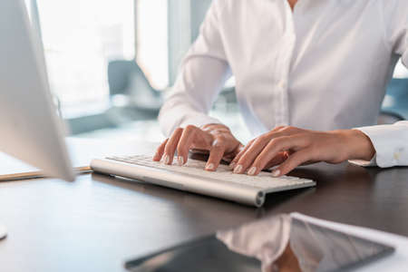 Woman office manager in white shirt sitting to a table with computer, hands on white keyboard...