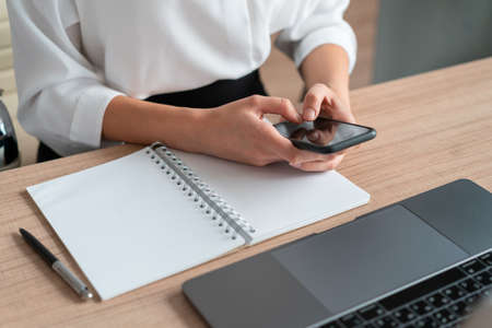 Top view of office manager, sitting to table with phone in hands and notebook and laptop on the...