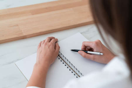 Top view of female hand holding a pen making notes in notebook, focus on blank paper list, woman office manager writing. Woman with notebook, no face. Concept of writingの写真素材