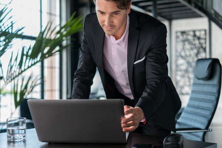 Young businessman wearing black suit, standing near table looking at the laptop. Office manager...