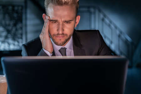 Tired businessman head in hand, working late at night in the office wearing black suit. Office manager working with laptop blurred background of dark office room. Concept of overworkの写真素材