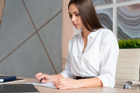 Office manager woman wearing white shirt, sitting to table with laptop and notebook, making notes...