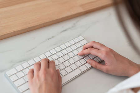 Top view of woman hands, fingers typing on white wireless keyboard lying on marble office table...