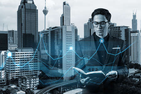 Young eastern handsome businessman in suit looking into appointment book to schedule the meeting and think about venture capital start up to estimate growth. Financial chart on Kuala Lumpurの写真素材