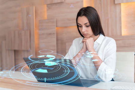 Attractive businesswoman in white shirt at workplace working with laptop to optimize development by implying new technologies in business process. Hi tech hologram over office backgroundの写真素材