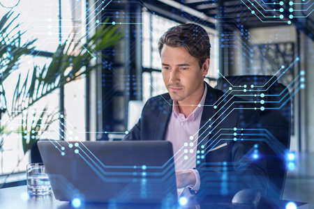 Handsome businessman in suit at workplace working with laptop to optimize development by implying new technologies in business process. Hi tech hologram over office backgroundの写真素材