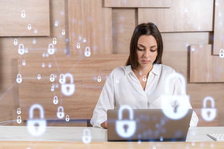 Attractive businesswoman in white shirt at workplace working with laptop to defend customer cyber security. Concept of clients information protection. Padlock hologram over office background.の写真素材