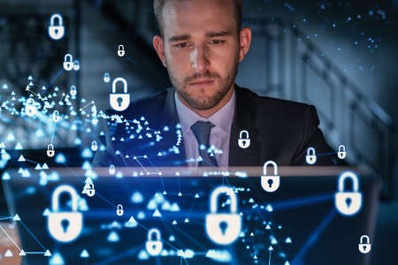 Handsome businessman in suit at workplace working with laptop to defend customer cyber security. Concept of clients information protection and brainstorm. Padlock hologram over office background.の写真素材