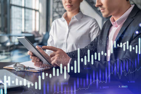 Businessman and businesswoman in formal wear working together to optimize trading strategy at corporate finance fund. Forex chart hologram over office background with panoramic windowsの写真素材