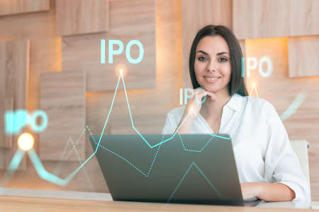 Attractive businesswoman in white shirt at workplace working with laptop to optimize IPO strategy at corporate fund. Financial chart hologram over office backgroundの写真素材