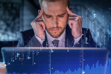 Handsome businessman in suit at workplace working with laptop to optimize trading strategy at corporate finance fund. Forex chart hologram over office backgroundの写真素材