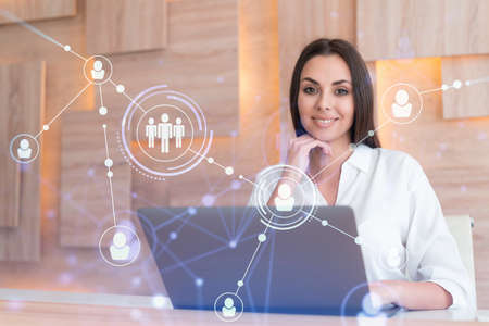 Attractive businesswoman in white shirt at workplace working with laptop to hire new employees for international business consulting. HR, social media hologram icons over office backgroundの写真素材