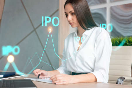 Portrait of attractive businesswoman in formal wear working with documents and thinking how to optimize IPO strategy. Financial chart hologram over modern office background. Go public concept.の写真素材