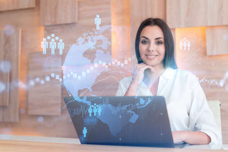 Attractive businesswoman in white shirt at workplace working with laptop to hire new employees for international business consulting. HR, social media hologram icons over office backgroundの写真素材