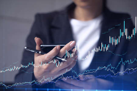 A woman trader holding in the hands a smart phone and researching stock market to proceed right investment solutions. Internet trading and wealth management concept. Hologram Forex chart.の写真素材