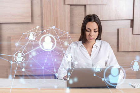 Attractive businesswoman in white shirt at workplace working with laptop to hire new employees for international business consulting. HR, social media hologram icons over office backgroundの写真素材